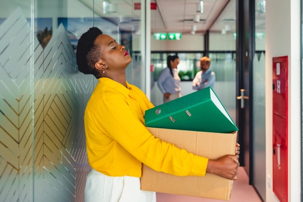A black woman with dark hair, wearing a yellow jumper and white bottoms leans against a patterned glass panel in a corridor whilst holding a box full of files,there are other people blurred in the background